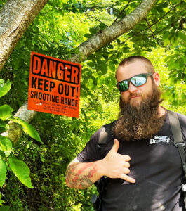 A bright orange Danger Shooting Range sign mounted on a tree with the owner of minuteman signs poseing next to it
